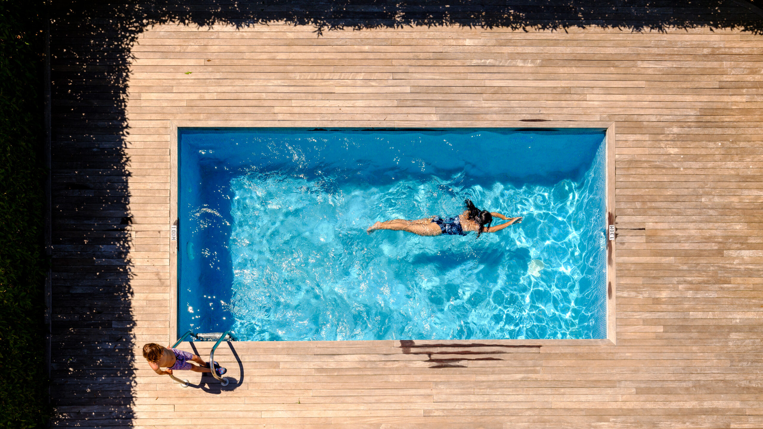 Drone view of anonymous little boy standing on wooden platform near young mother swimming in pool on sunny day during summer vacation