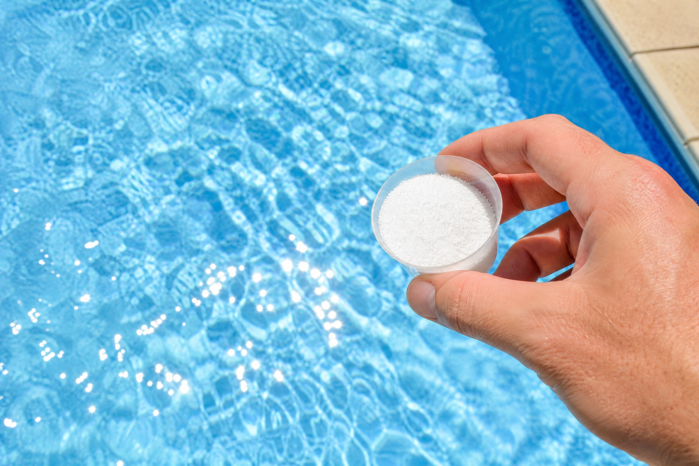 Stabilisant de piscine : main tenant un doseur rempli de granulés blancs au-dessus d'une eau bleue claire
